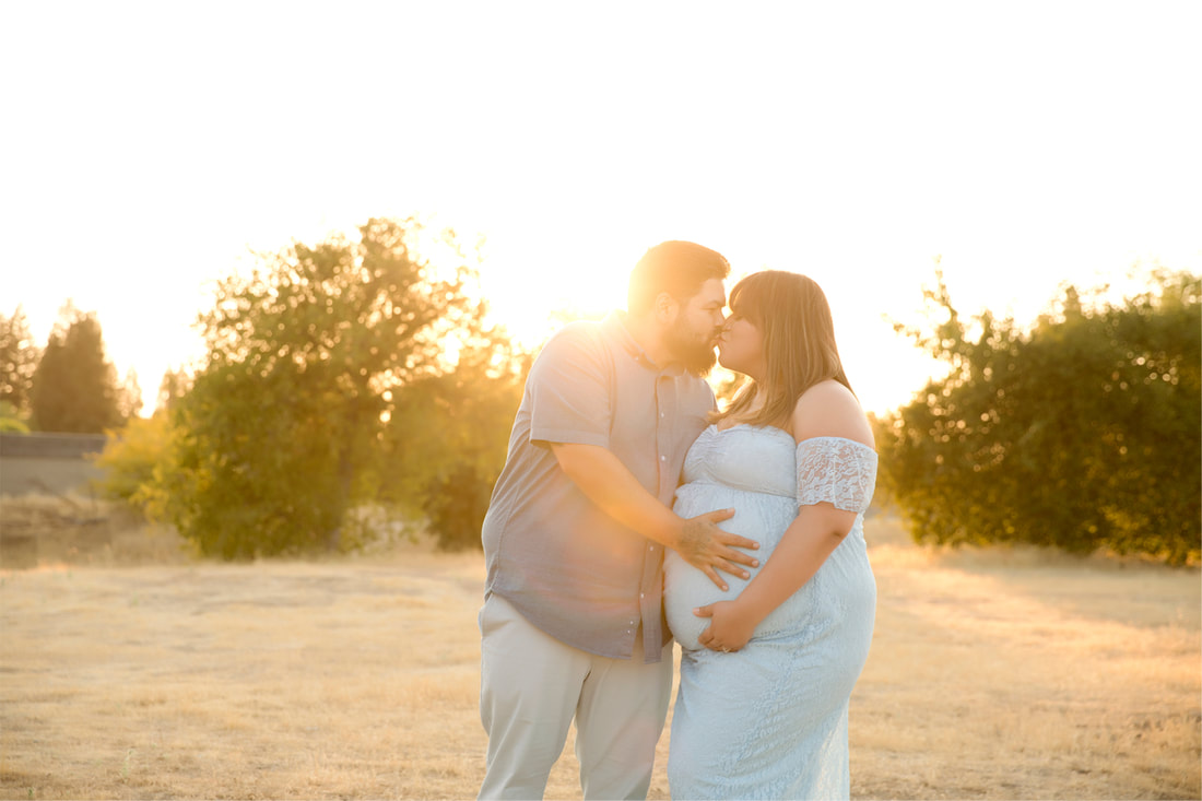 Man kissing pregnant woman in blue, holding her belly. Greenery and sunset behind them